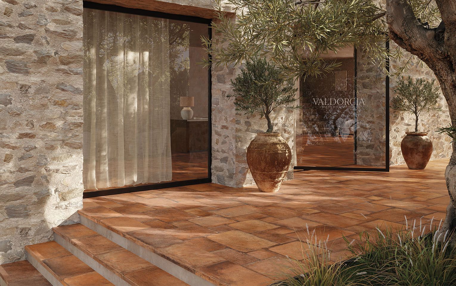 Charming outdoor terrace featuring Icotto Cotto tiles in warm terracotta hues, seamlessly blending with a rustic stone facade. Large ceramic pots with olive trees enhance the Mediterranean ambiance, while sheer curtains softly filter the natural light from inside the stone house. The image captures the timeless elegance of Tuscan-inspired design, where the earthy tones of the terracotta tiles complement the rugged beauty of the stone walls, creating a serene and inviting outdoor living space.