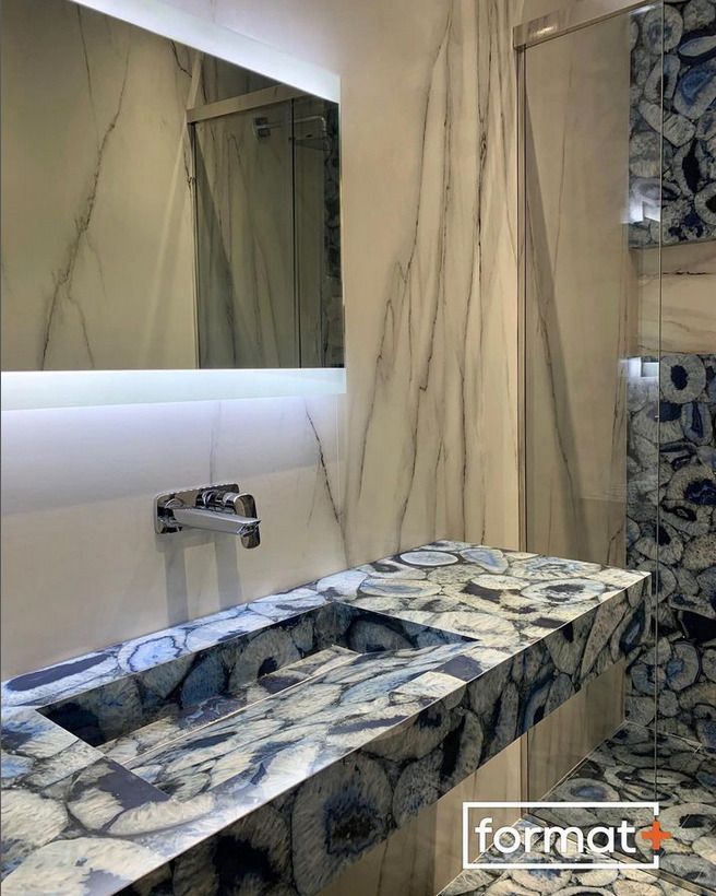 A bathroom interior featuring a Format+ branded stone sink with bold blue and white patterns, accompanied by a backlit mirror and matching stone shower details.