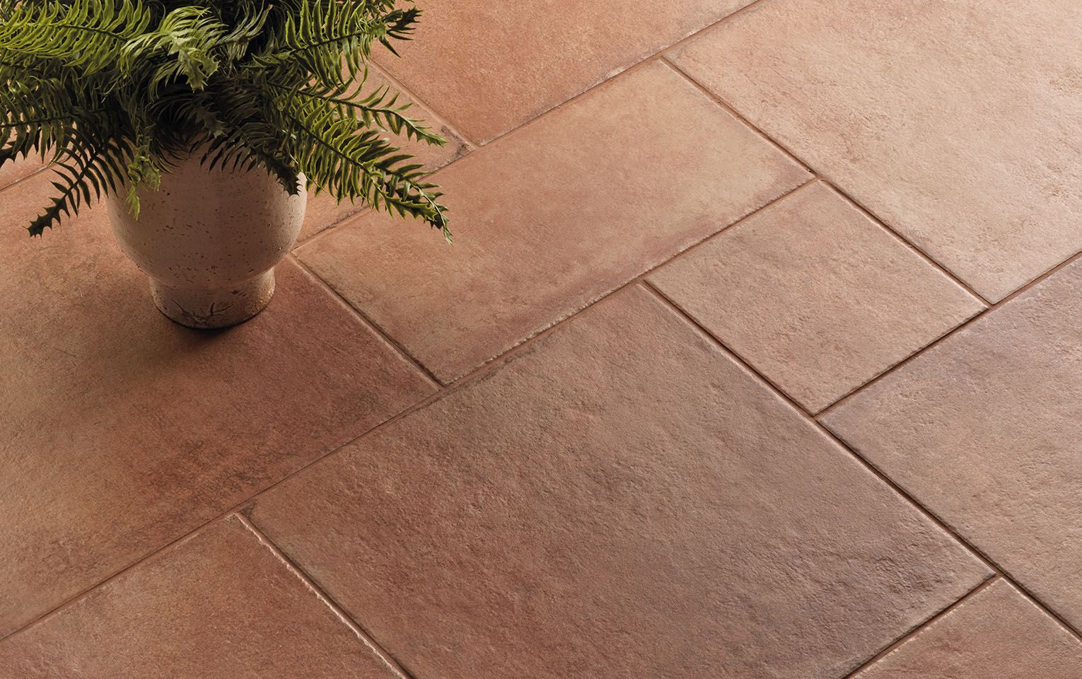 Close-up of Icotto Aurora tiles in soft, warm terracotta hues, featuring a natural, textured surface that adds depth and character to the flooring. A ceramic vase with lush green ferns sits on the tiles, creating a harmonious blend of nature and rustic design. The image highlights the versatility and timeless appeal of these tiles, perfect for adding warmth and elegance to both indoor and outdoor spaces.