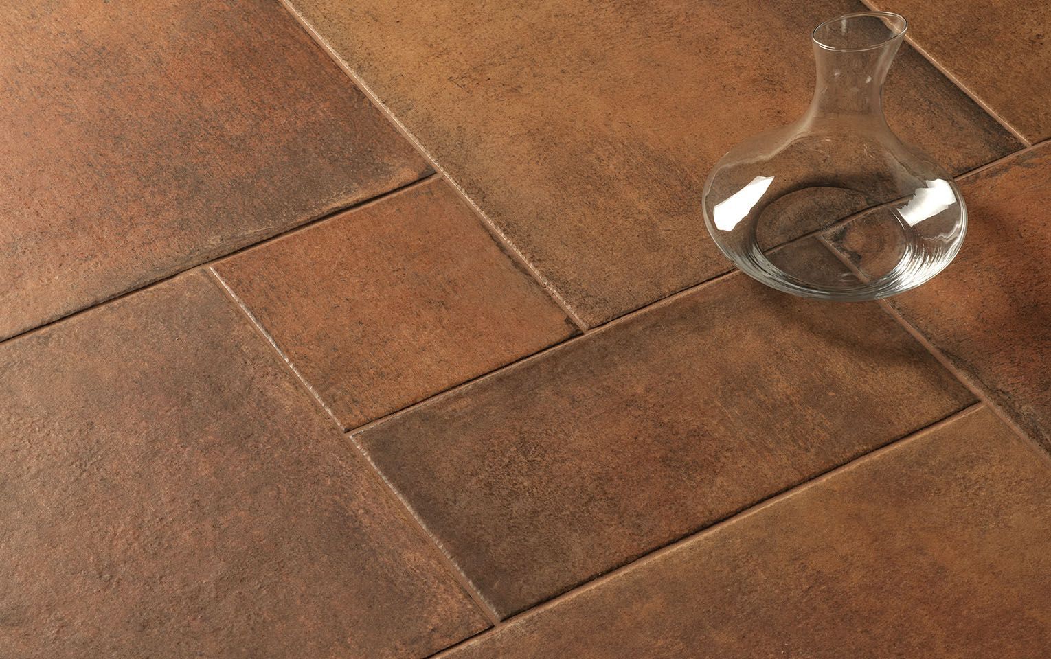 Close-up of Icotto Cotto tiles in warm, rich earthy tones, highlighting the natural texture and variation of terracotta-inspired surfaces. A clear glass carafe placed on the tiles adds a modern touch, contrasting with the rustic aesthetic. The image emphasizes the durability and timeless beauty of the tiles, making them ideal for creating a classic yet contemporary atmosphere in any living space