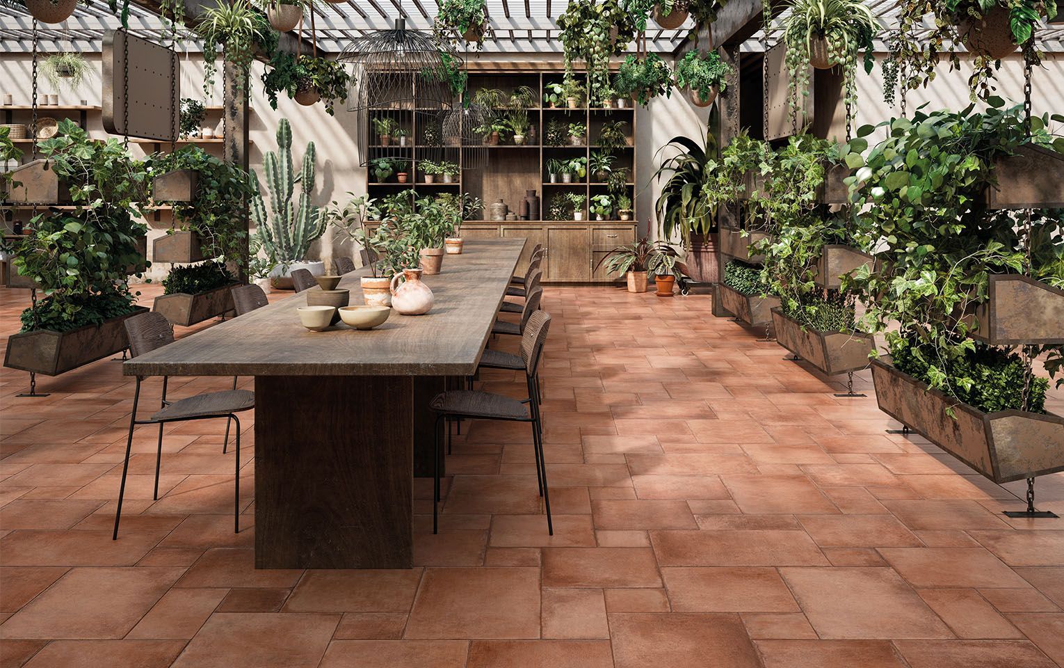 Inviting indoor garden space featuring Icotto Aurora tiles in warm terracotta tones, complemented by an abundance of lush greenery and hanging plants. A large wooden table with rustic chairs serves as the centerpiece, surrounded by a variety of potted plants, cacti, and unique planters. The combination of natural elements and earthy tile flooring creates a serene and organic environment, perfect for gatherings or a peaceful retreat within a greenhouse-like setting