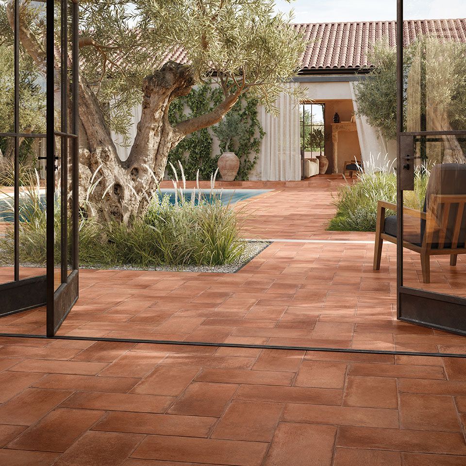 Terracotta floor tiles in an outdoor patio space with Mediterranean landscaping. An ancient olive tree and lush greenery surround the area, leading to a cozy pool.