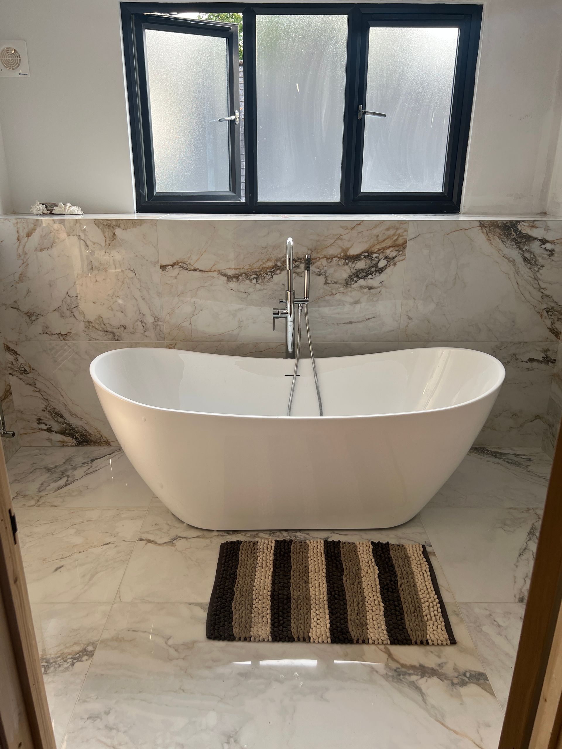A luxurious bathroom featuring white marble tiles, a freestanding bathtub, and a large window with frosted glass. A striped bathmat is placed on the floor.