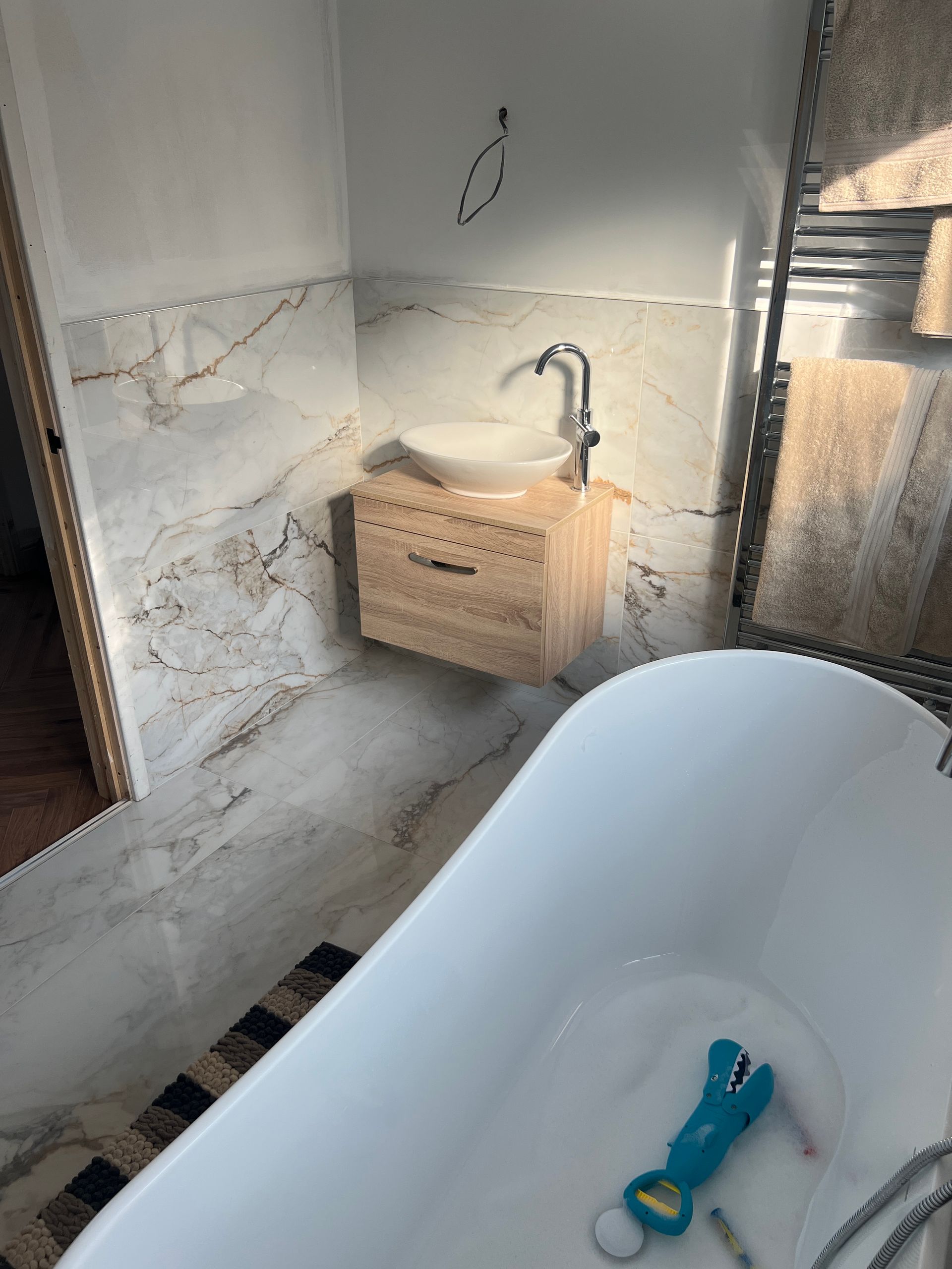 A modern bathroom sink with a light wooden vanity, placed against glossy marble wall tiles. A freestanding bathtub is visible in the foreground.