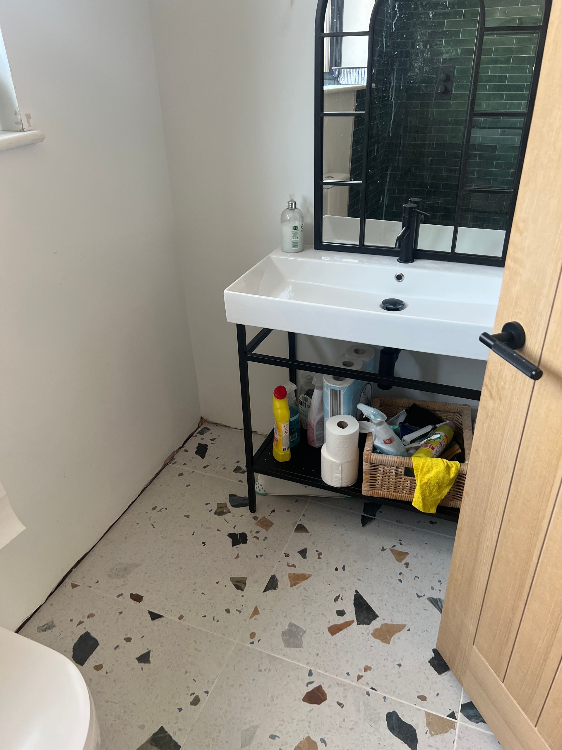A small bathroom sink and vanity area featuring a black-framed mirror, terrazzo floor tiles, and a window providing natural light.