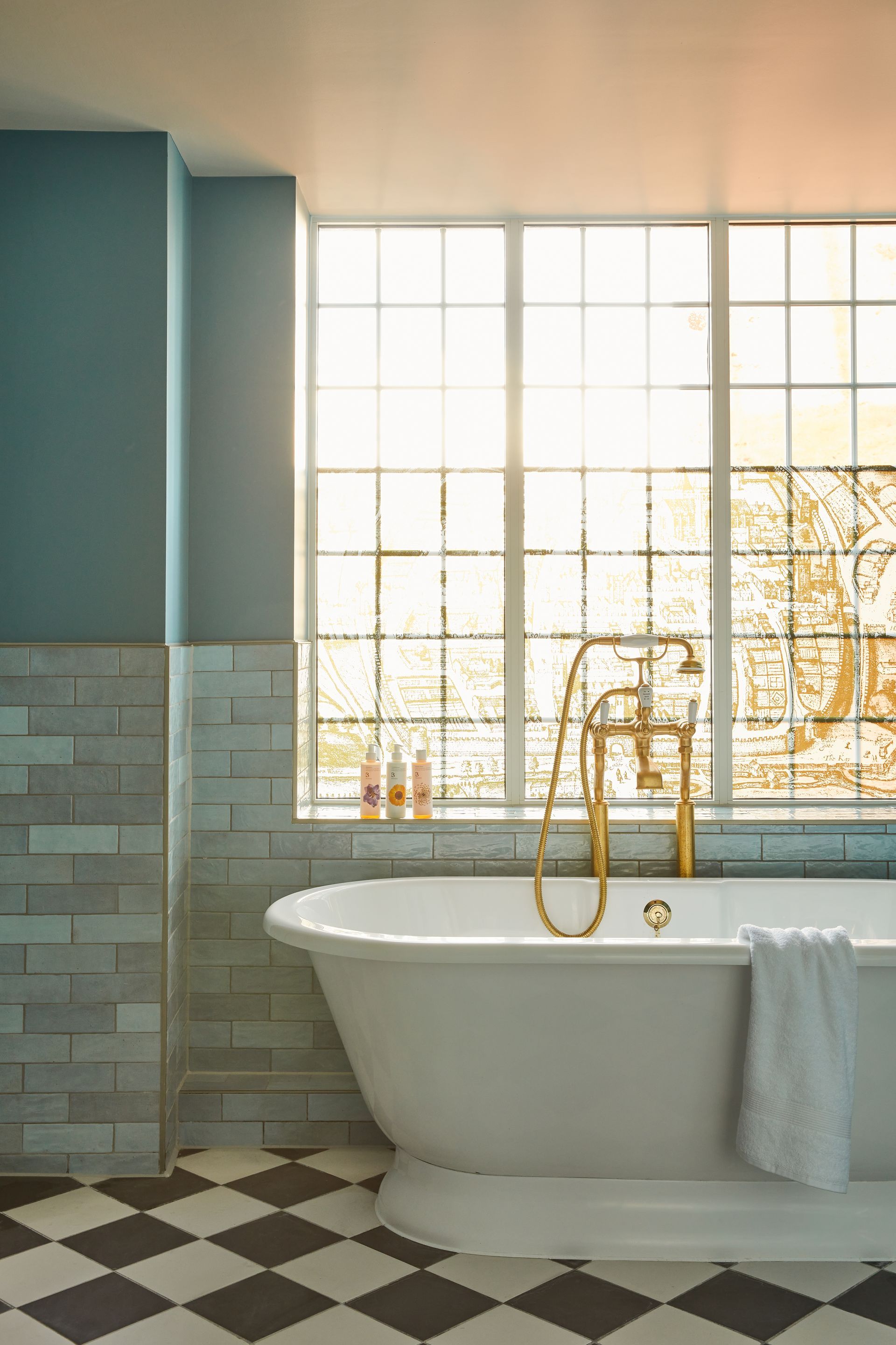 Golden bath tap Infront of glass pains with sunlight shining through, blue lyon tiles on the wall