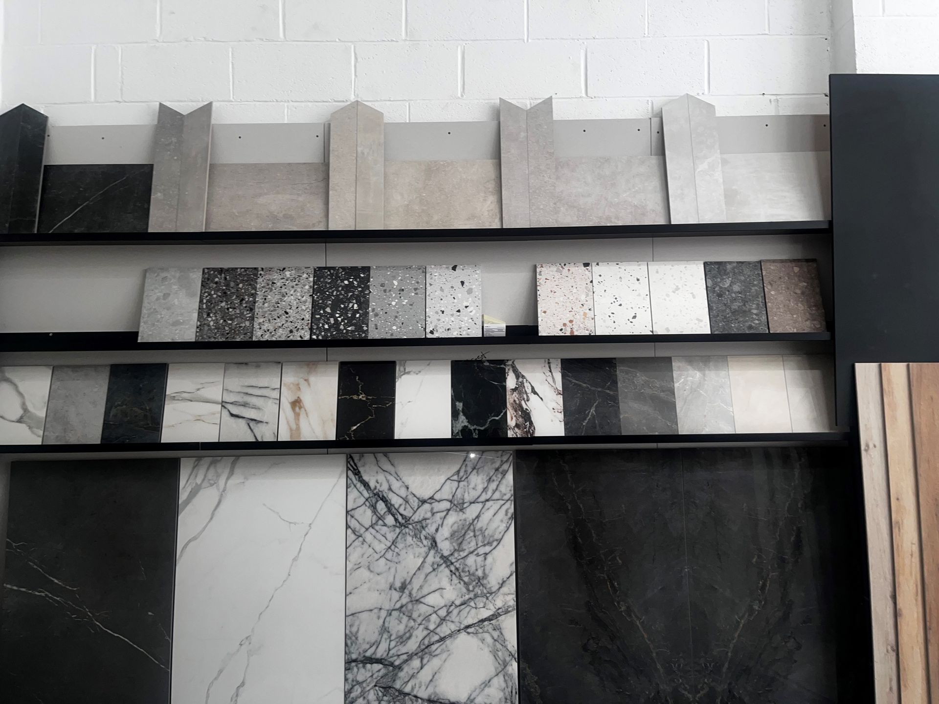 A display of colorful, textured tiles arranged on shelves in a tile showroom. The display features a variety of tile patterns, textures, and colors, from pastel pinks and greens to terrazzo and wood effects.