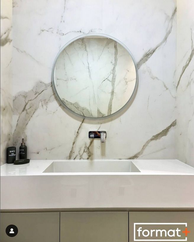 A clean and elegant bathroom featuring a circular mirror, white marble backsplash, and a simple rectangular sink with subtle gold accents.