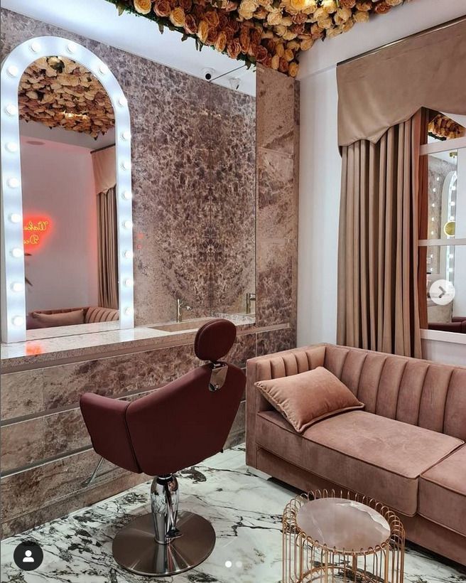 A glamorous salon vanity featuring a marble countertop and plush pink seating, accented by a mirror with lightbulbs and floral decor.