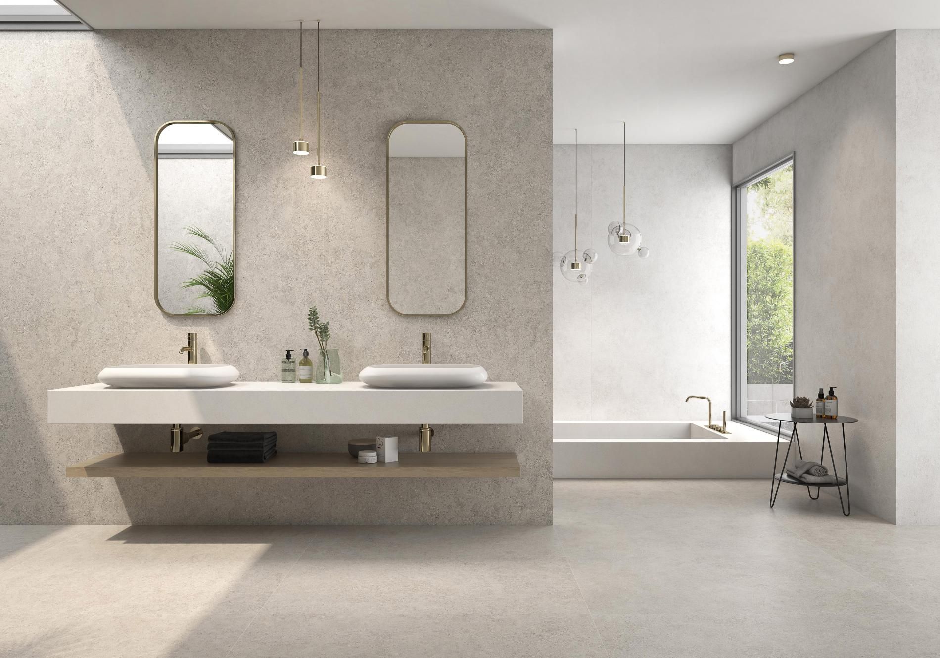 A spacious bathroom with a minimalist double vanity. Light beige stone-effect tiles cover the walls and floor. The space is illuminated with soft pendant lights, creating a calming atmosphere. Large windows provide ample natural light.