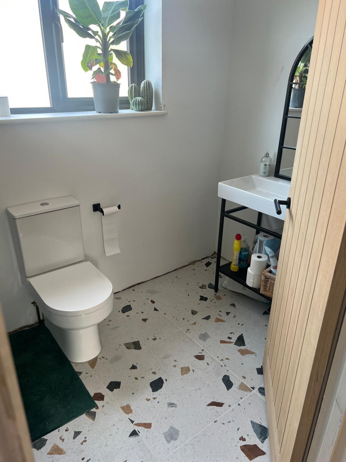 A minimalistic bathroom with a white toilet, terrazzo floor tiles, and a potted plant on the windowsill, next to a small sink and vanity.