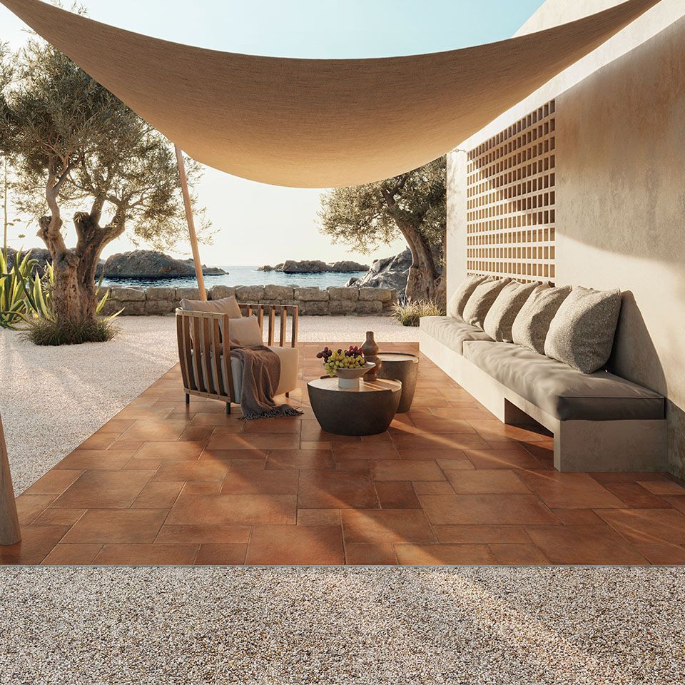 Serene outdoor lounge area featuring Icotto Cotto tiles in warm terracotta tones, set against a backdrop of the sea and rocky shoreline. The space is shaded by a large sail canopy, with a comfortable built-in bench adorned with soft cushions and a modern wooden armchair. A pair of round coffee tables with decorative items adds to the cozy ambiance. Olive trees and natural stone elements enhance the Mediterranean vibe, creating an inviting and peaceful retreat by the water