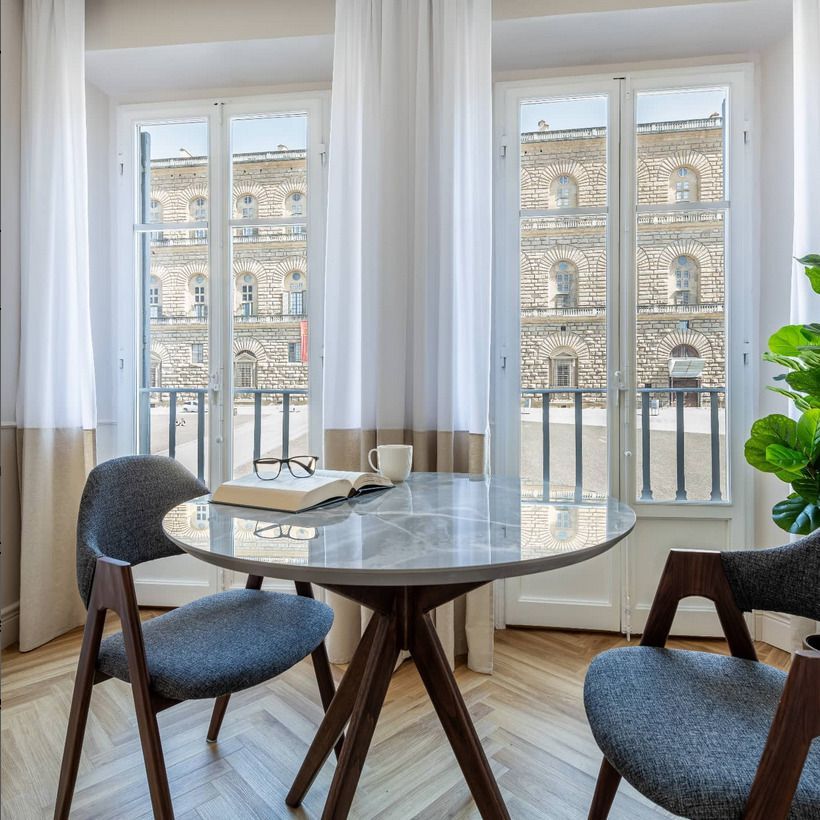 A small table and chairs set near large windows, offering a bright, airy ambiance with a view of an old stone building.