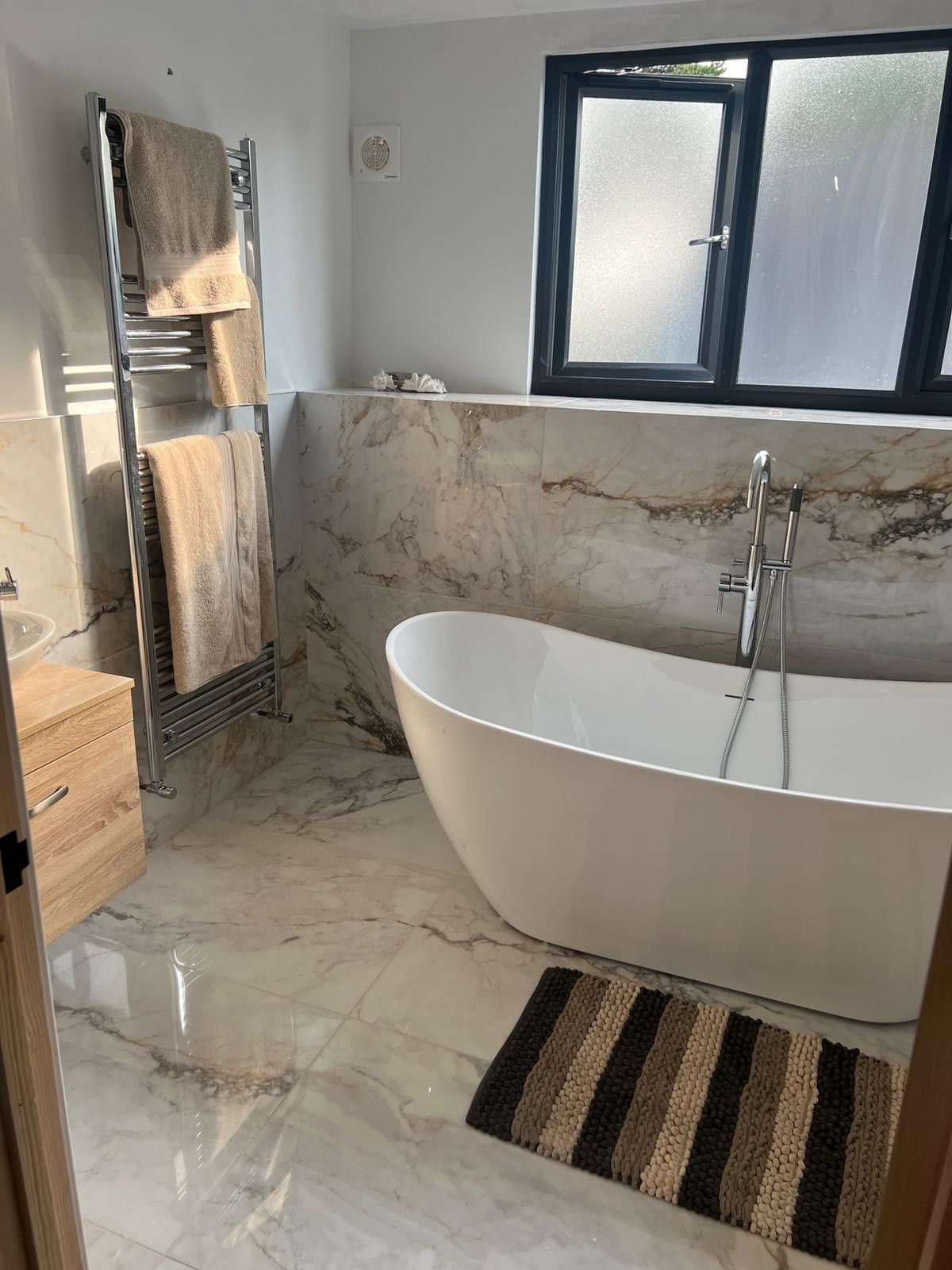 A close-up of a freestanding white bathtub with a nearby chrome towel rack in a marble-tiled bathroom. The walls and floors are both made of glossy marble tiles.