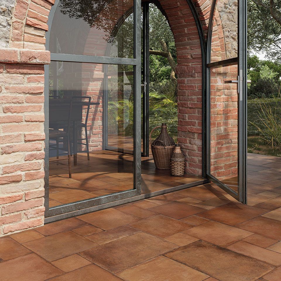 Charming patio entrance featuring Icotto Cotto tiles in warm terracotta tones, seamlessly blending with the rustic brick arches and stone facade. The floor-to-ceiling glass doors open to reveal a cozy interior dining area, while woven baskets and natural greenery add to the rustic Mediterranean ambiance. The rich, earthy tiles enhance the traditional architectural elements, creating a welcoming and timeless outdoor-to-indoor transition