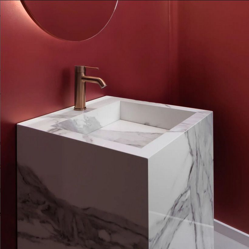 A minimalist white marble pedestal sink against a bold red wall, with a bronze faucet.