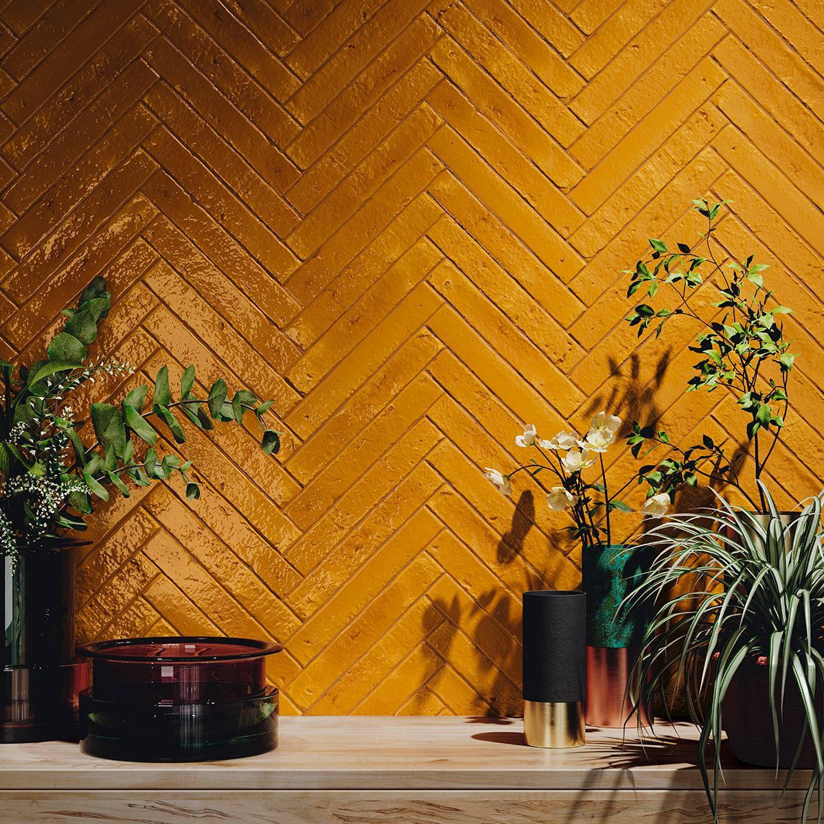 Interior wall featuring mustard-coloured distressed brick-effect tiles arranged in a herringbone pattern, adorned with modern decorative plants and vases on a wooden shelf, creating a bold and stylish design statement