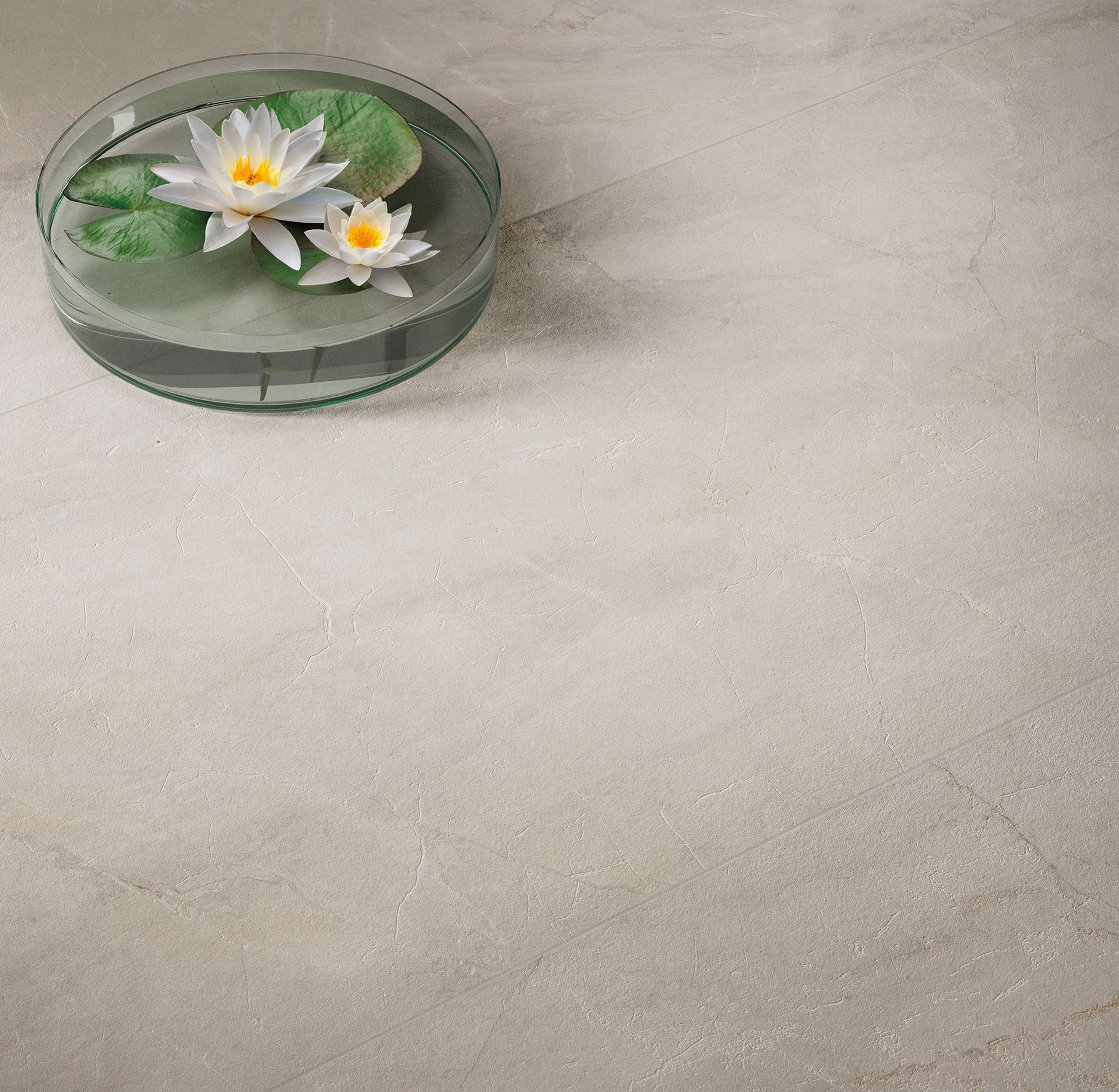 A top-down view of beige marble-effect tiles, adorned with a decorative bowl containing water lilies. The delicate flower arrangement enhances the serene and natural aesthetic of the space.