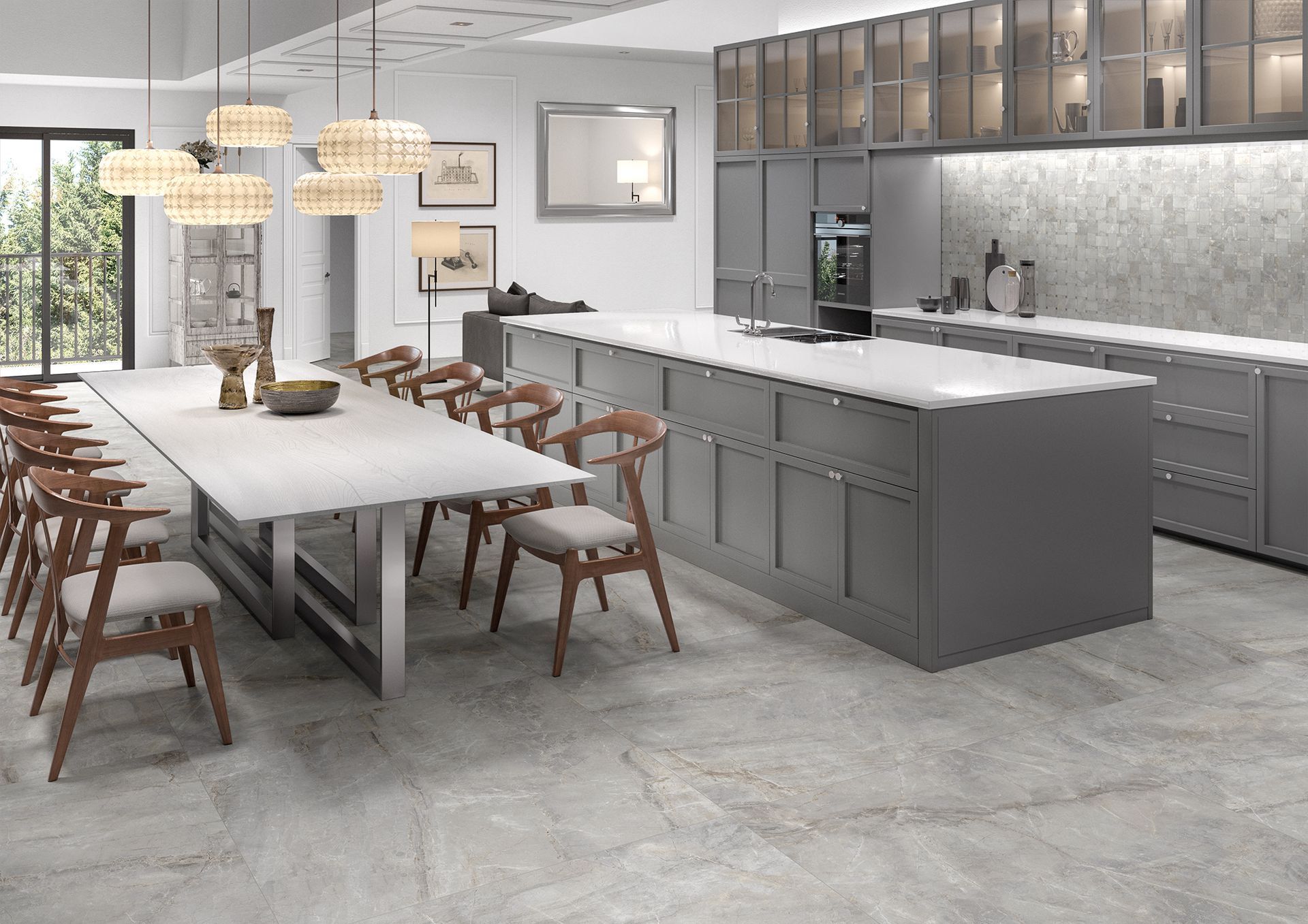 A bright, open kitchen featuring light gray stone floor tiles. The kitchen is equipped with a large white island countertop, light gray cabinetry, and wooden chairs around a dining table. Modern pendant lights hang above, adding a touch of warmth.