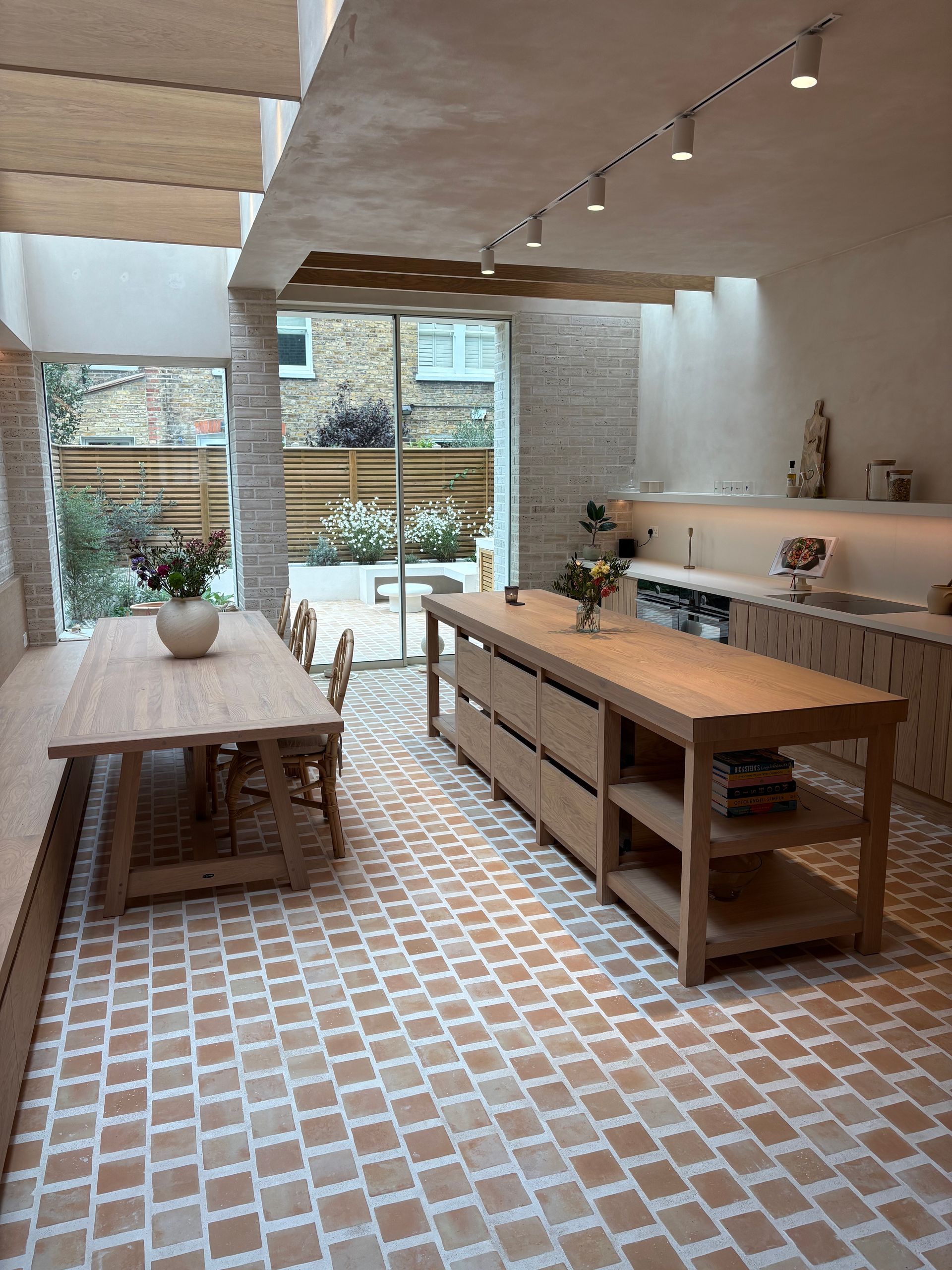 Warm terracotta tiles with a green accent border in a Mediterranean-style entrance hallway. The room features arched ceilings, minimalist furniture, and potted plants.