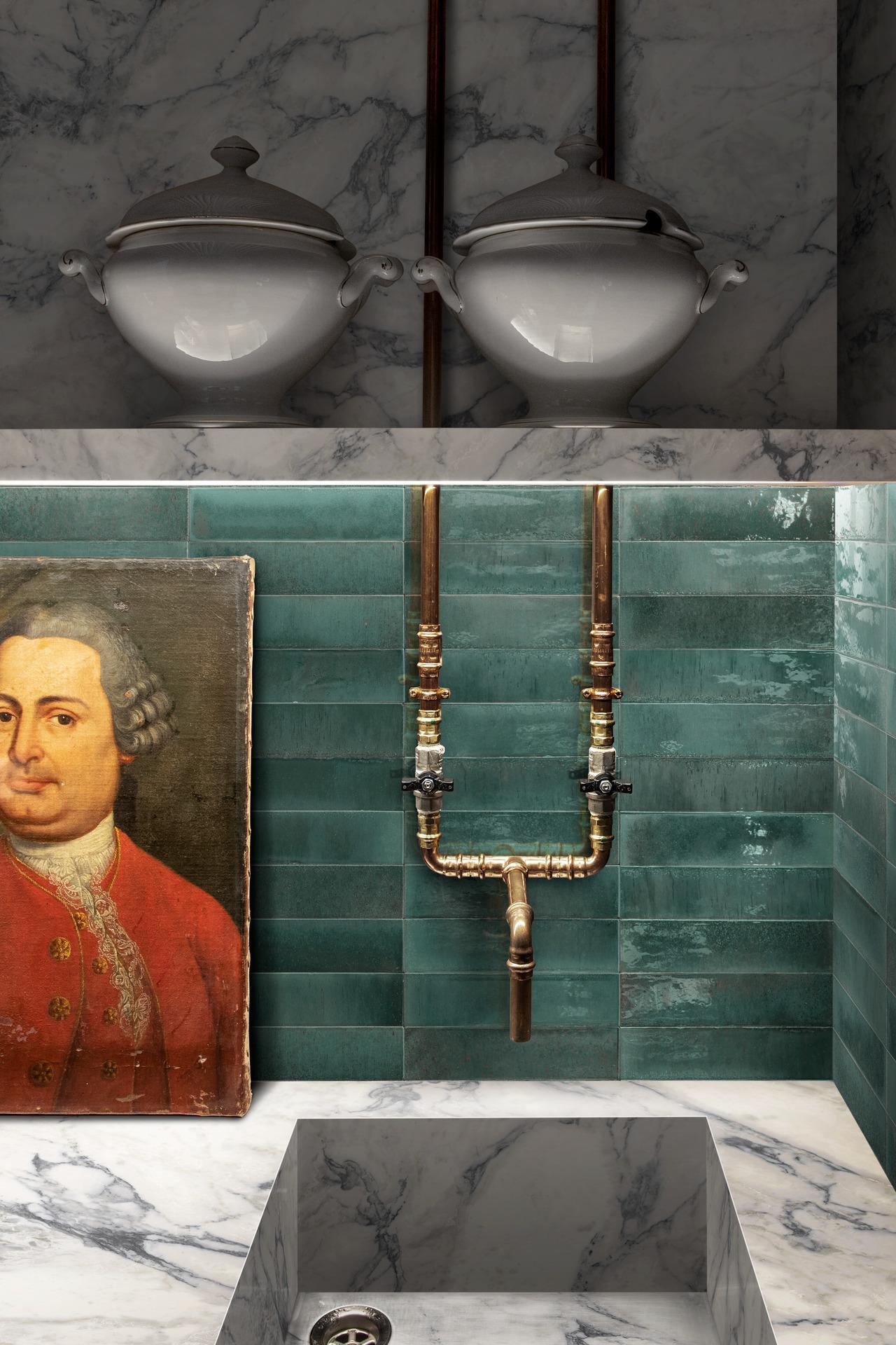 Eclectic bathroom design featuring glossy green zellige-effect tiles, a marble countertop and sink, exposed copper piping, and a classical portrait, creating a unique blend of vintage and modern elements.