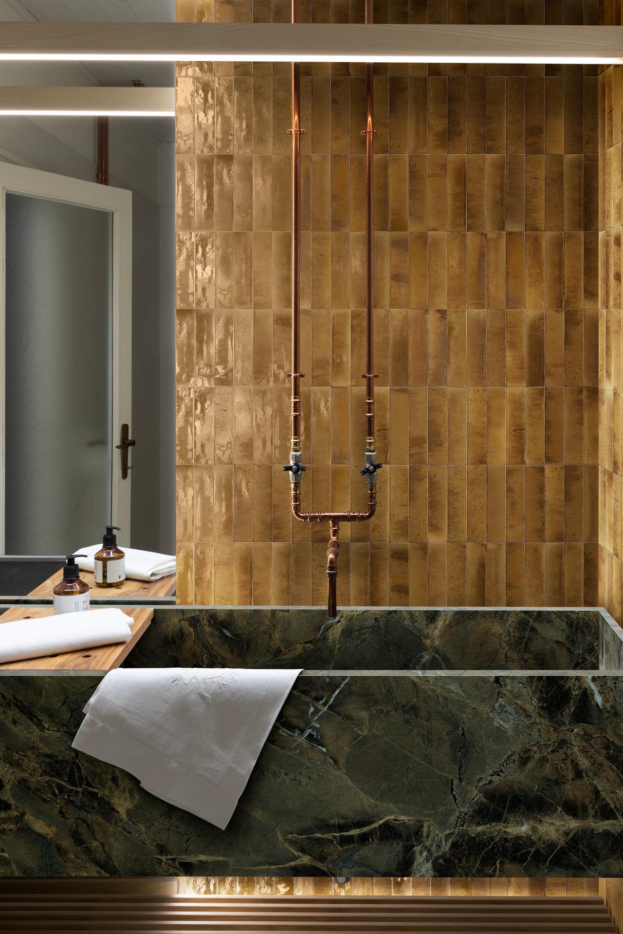 Luxurious bathroom featuring a deep green marble bathtub with copper piping and walls adorned with glossy golden zellige-effect tiles, creating an opulent and contemporary design.