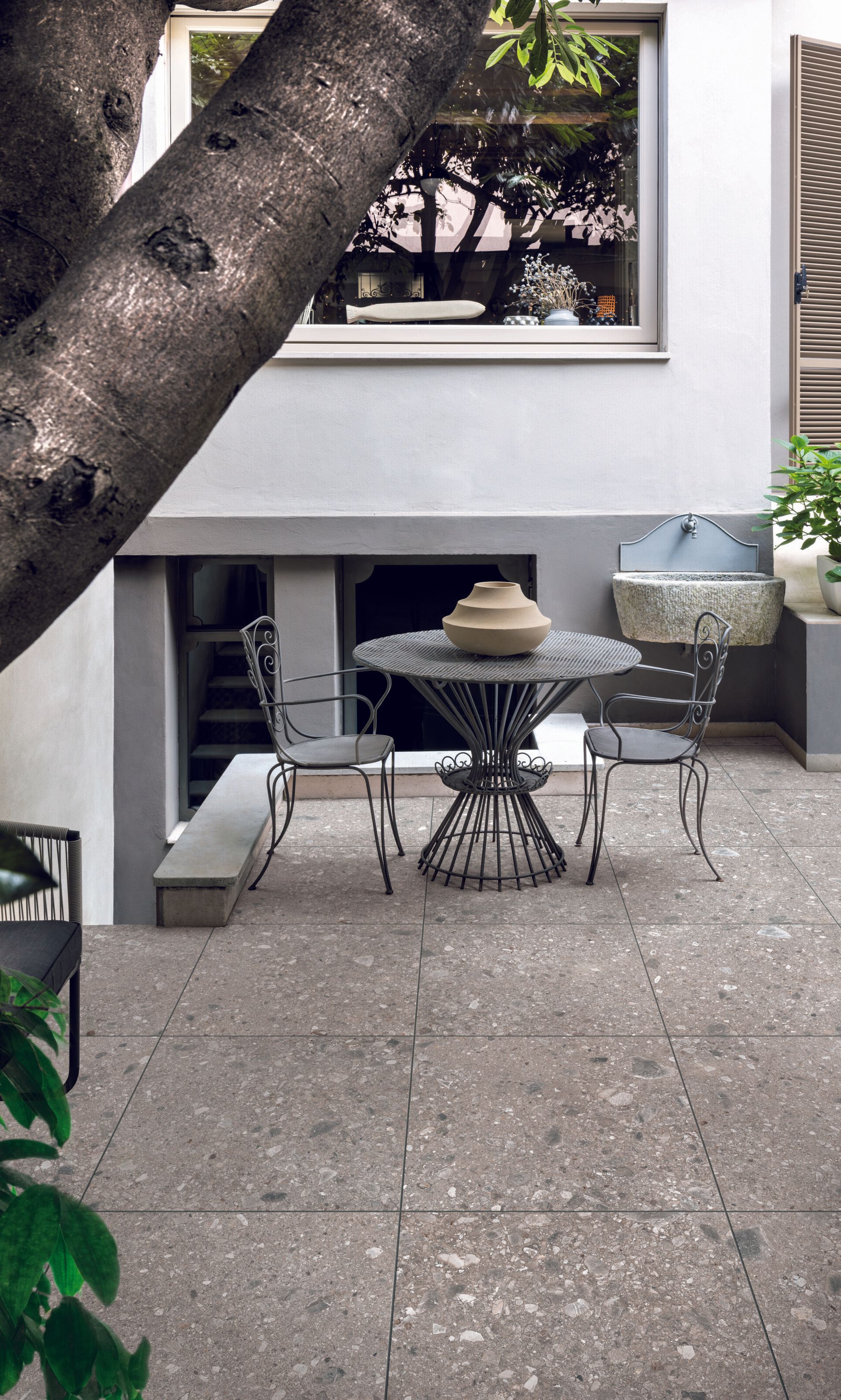 Small courtyard with terrazzo-style tiles and classic wrought iron table and chairs. Tree and lush greenery enhance the cozy garden setting.