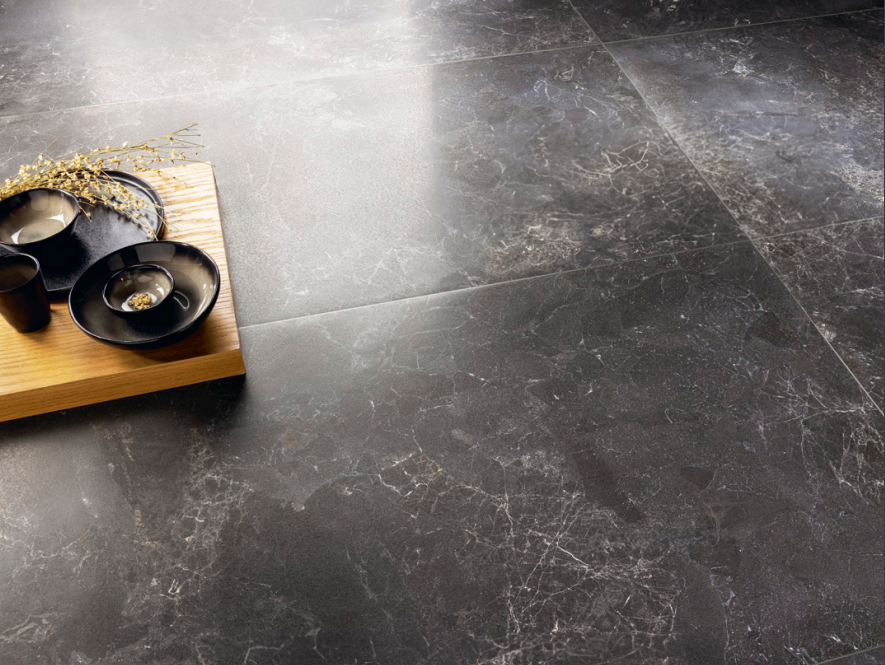 Close-up view of dark marble floor tiles with prominent veining. A wooden tray holds a set of black ceramic dishes and a small decorative branch, highlighting the rich texture and elegant appearance of the tiles. The lighting accentuates the natural patterns and polished surface.