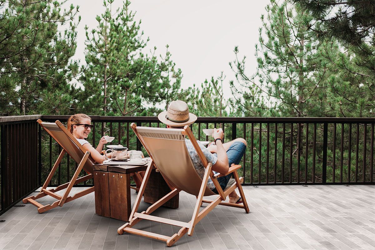 Two people relaxing on a balcony surrounded by pine trees. The space is finished with grey rectangular outdoor tiles and wooden lounge chairs.
