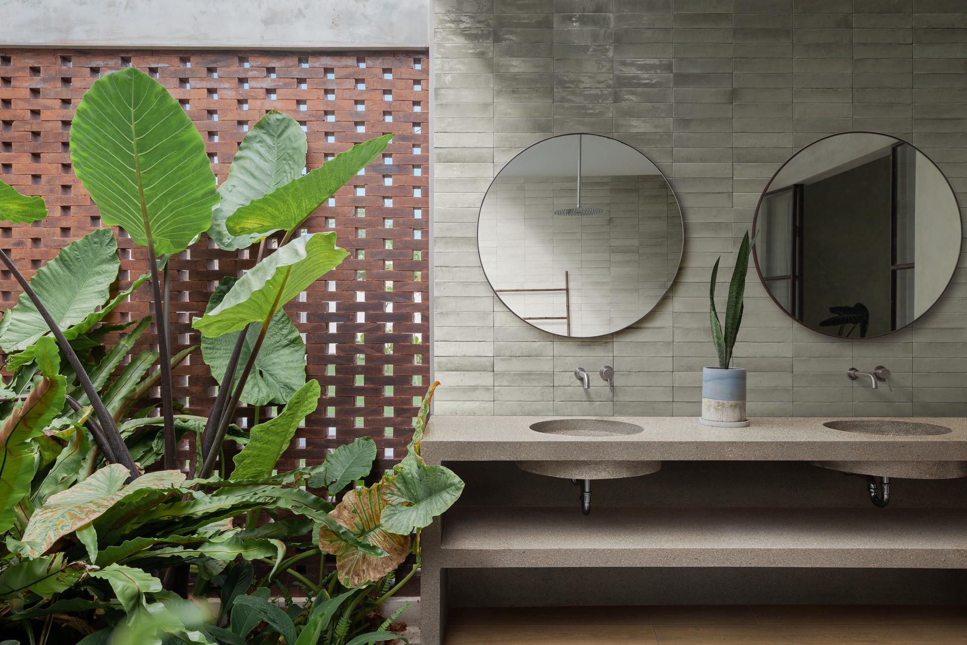 Modern bathroom featuring a dual sink vanity with a terrazzo countertop, glossy gray zellige-effect tiles, round mirrors, and large tropical plants next to a brick screen wall, blending indoor and outdoor elements
