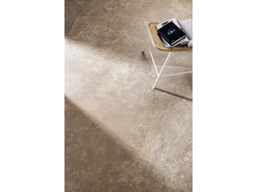 Overhead view of a beige marble floor tile with subtle veining, creating a natural and elegant look. A wicker and metal chair with a few magazines placed on it is visible at the top of the image, adding a casual and cosy element to the scene. The soft lighting enhances the texture and colour variations of the tiles