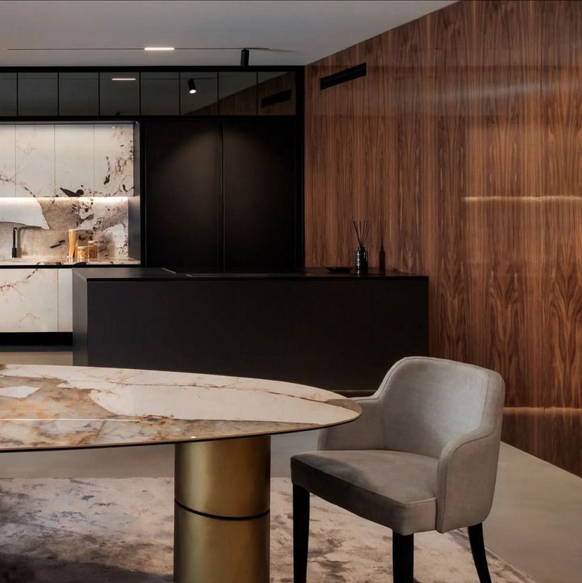 A contemporary dining room showcasing a sleek oval marble dining table with gold cylindrical legs. The room is complemented by black cabinetry and a marble backsplash in the kitchen area.