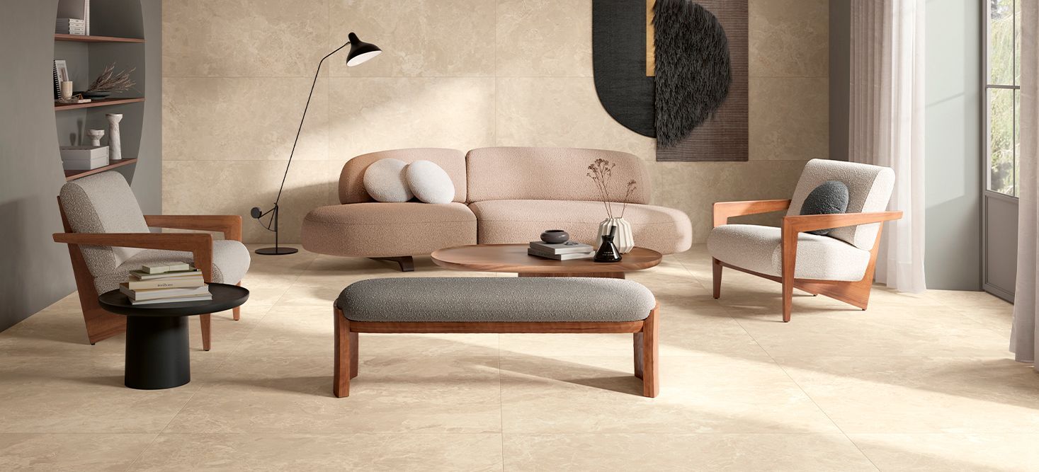 Modern living room with light beige marble flooring and neutral-coloured furniture. The room features a curved beige sofa, two wooden armchairs with light grey cushions, and a matching bench. A round black side table holds a stack of books, while a wooden coffee table displays decorative items. The room has a minimalistic design with a floor lamp, a wall-mounted artwork, and built-in shelves, creating a cozy and stylish atmosphere