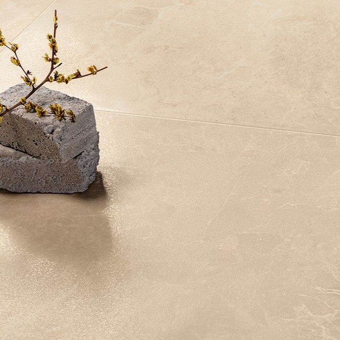 Close-up view of a light beige marble floor tile with subtle veining. A decorative element featuring a grey stone block with a branch of small yellow flowers rests on the tile, showcasing the smooth and polished surface of the flooring