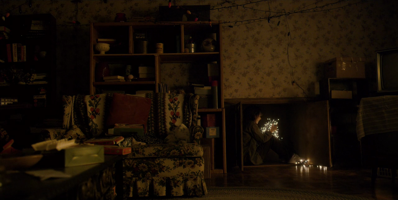 Dark interior shot of the byers livingroom with Joyce byers sat inside storage space clasping lit string lights.