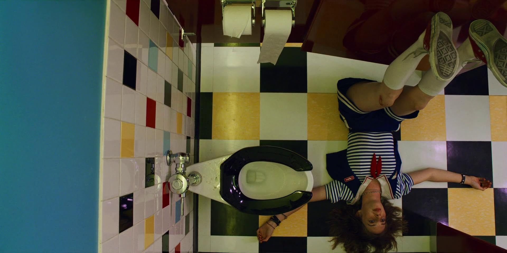 Robin from stranger things laying carelessly with her feet at a 90 degree angle against a red door, laying in the mall restroom, which is adorned in colourful square tiles.