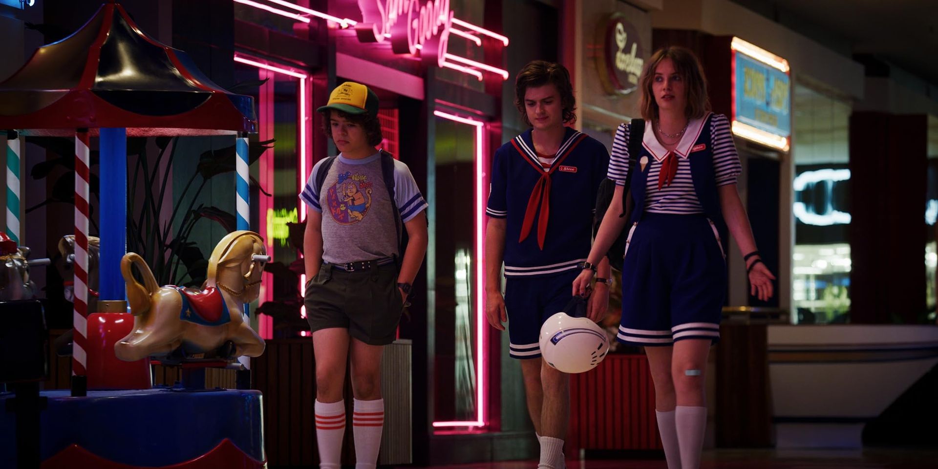 from left to right, Dustin, Steve and Robin walk through the mall in their scoops ahoy uniforms after closing time.