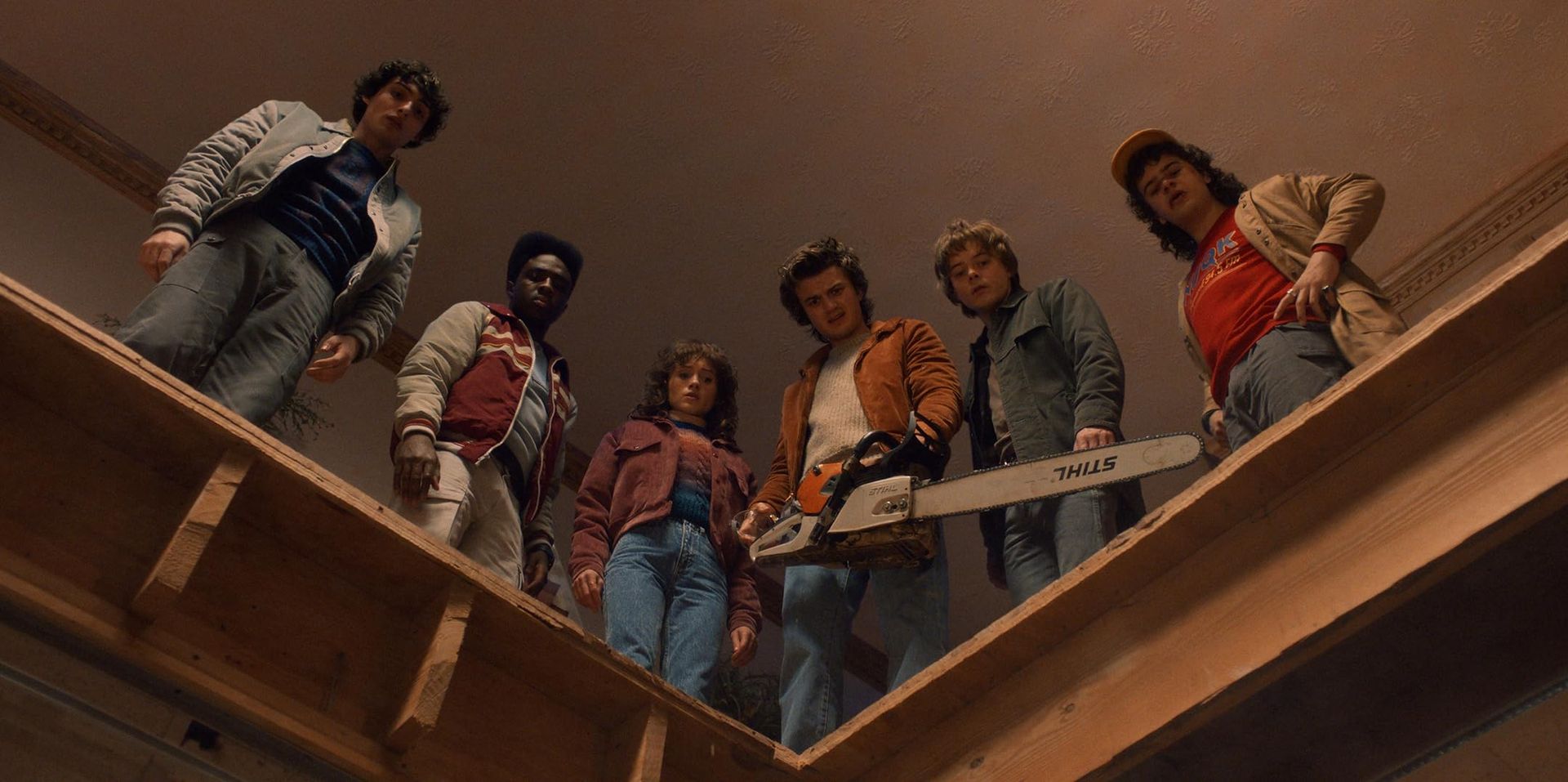 from left to right, Mike, Lucas, Nancy, Steve, Jonathan and Dustin. All looking down through a hole in the floor. steve is holding a chainsaw.