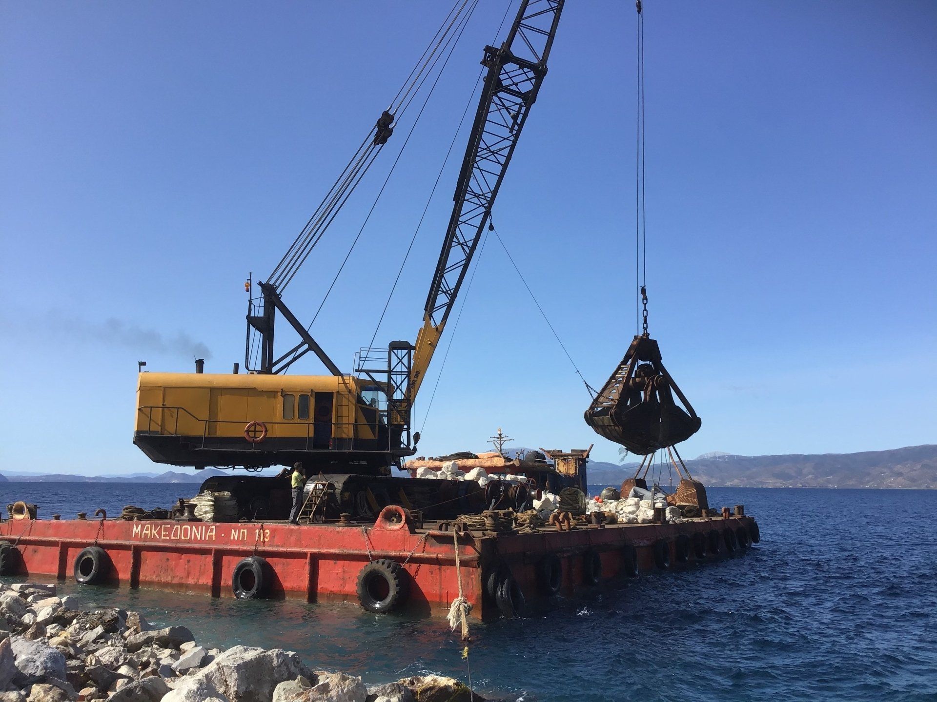 Sea crane-barge delivering stone to repair Kamini Harbour on Hydra Island Greece.