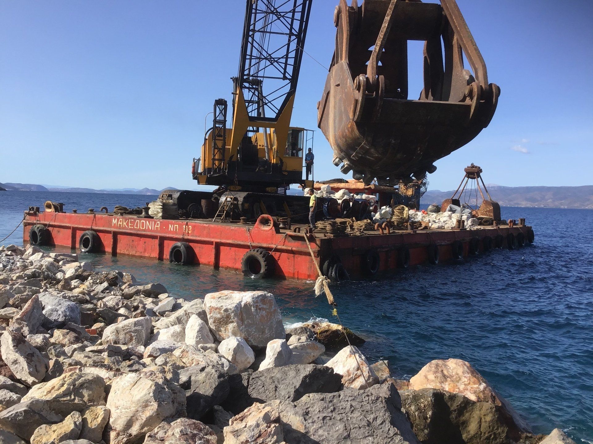 Sea crane-barge delivering stone to repair Kamini Harbour on Hydra Island Greece.