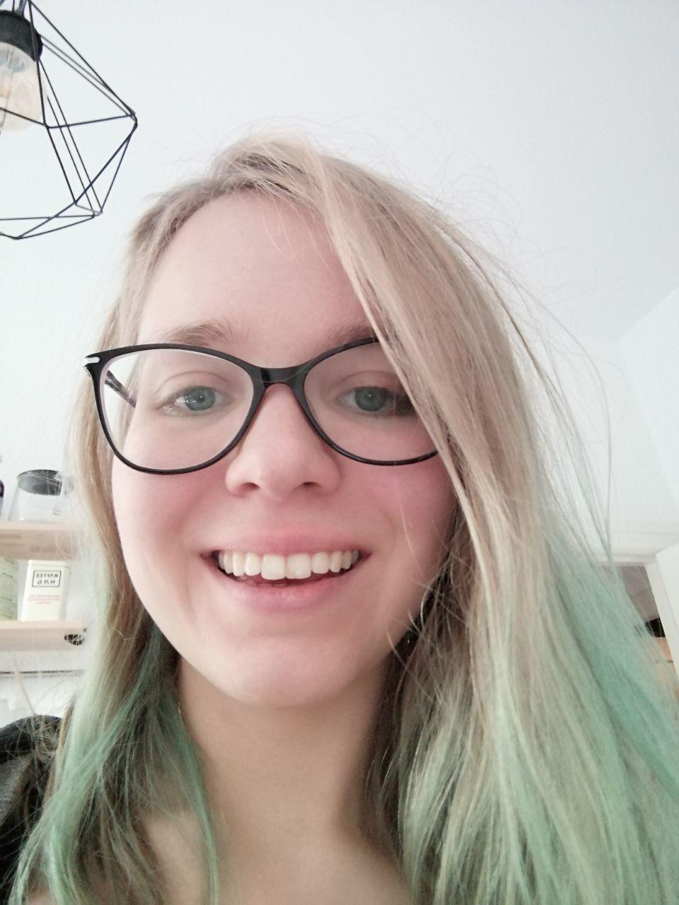 Foto von Swantje Niemann, das fast ganz von ihrem lächelnden Gesicht eingenommen wird. Sie trägt eine Brille und hat blonde Haare, an deren Spitzen man schwach eine fast herausgewaschene, blau-grüne Tönung sieht. Im Hintergrund sind ein Regal und eine Lampe zu sehen.