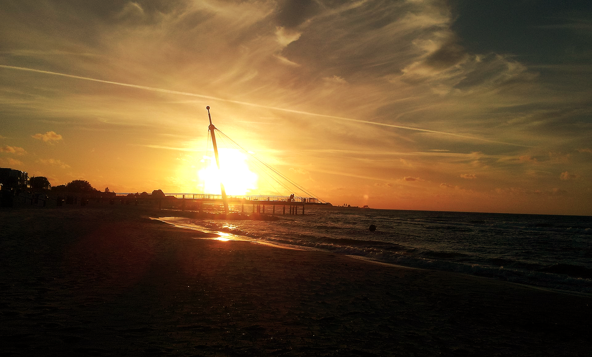 Abendlicher Blick auf die Flunder in Hohwacht. Ein rot-oranger Sonnenuntergang, vermischt mit verlaufenden Wolken. Ein paar Menschen stehen auf der Flunder und geniessen diesen wunderbaren Augenblick. Im Wasser spiegelt sich die Sonne und der Schatten der scheinbar schwebenden Aussichtsplattform