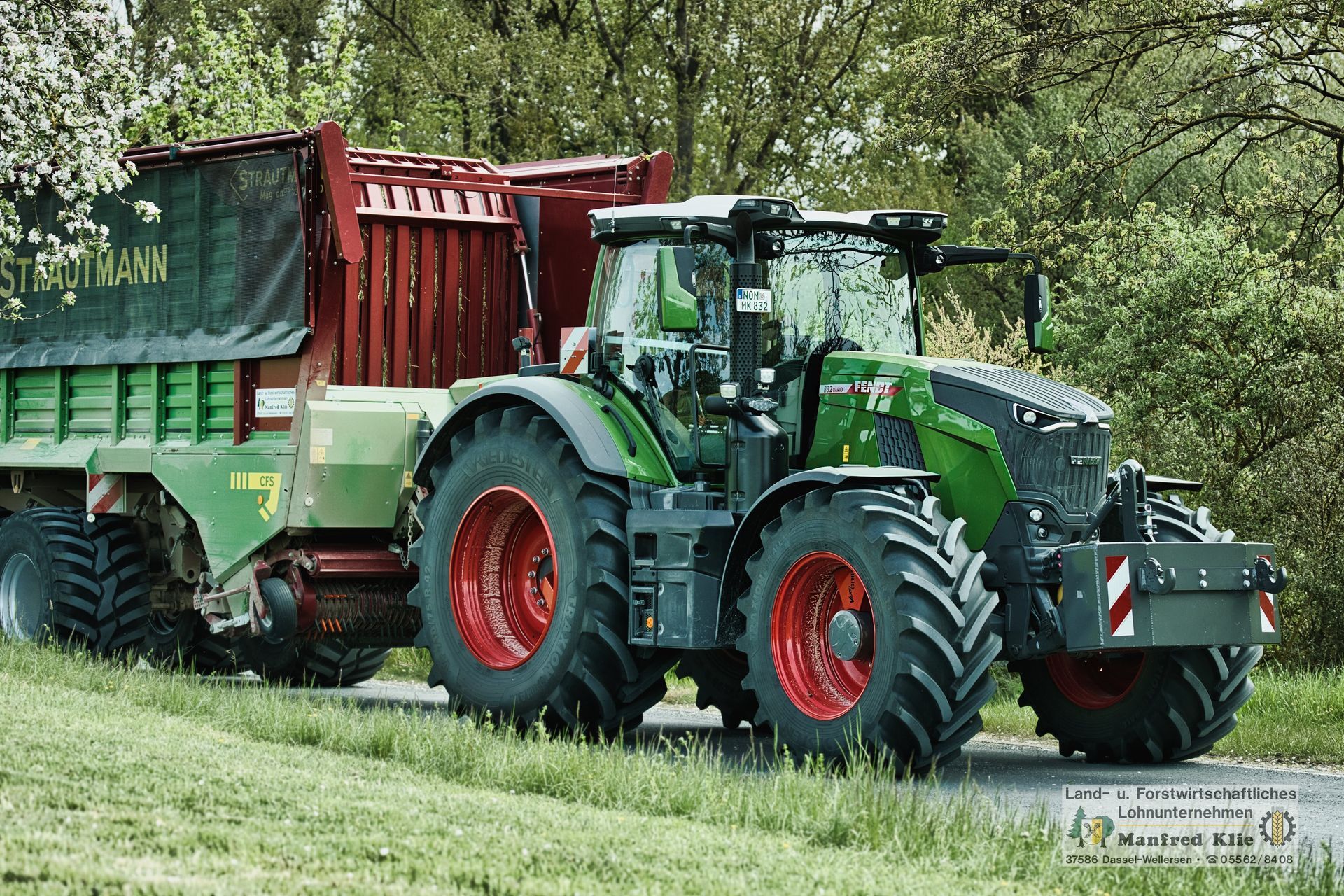 Moderner grüner Fendt 832 Traktor mit roten Felgen fährt auf einer ländlichen Straße und zieht einen großen Strautmann-Ladewagen, umgeben von Bäumen und Wiesen.