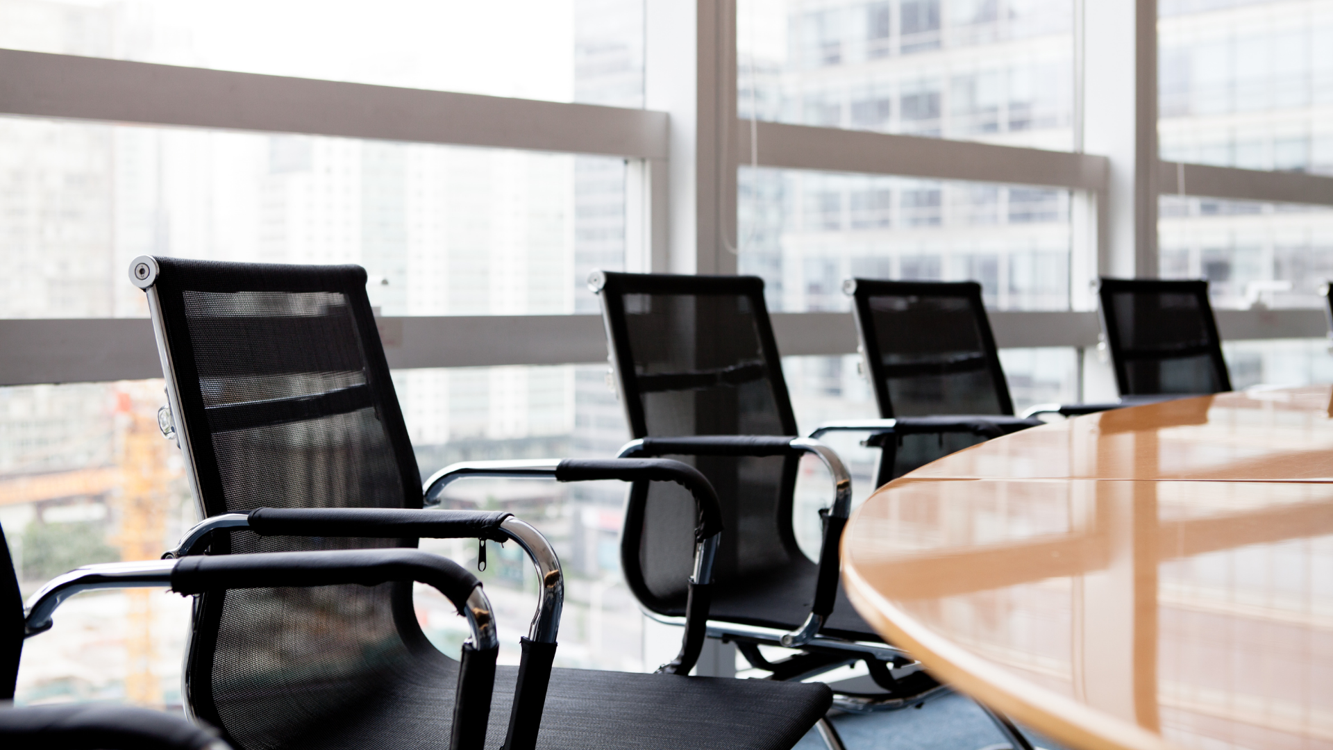 Schwarze Bürostühle an einem Holztisch vor einer Fensterfront. Das Bild symbolisiert einen Besprechungstisch, an dem der Beirat tagt.