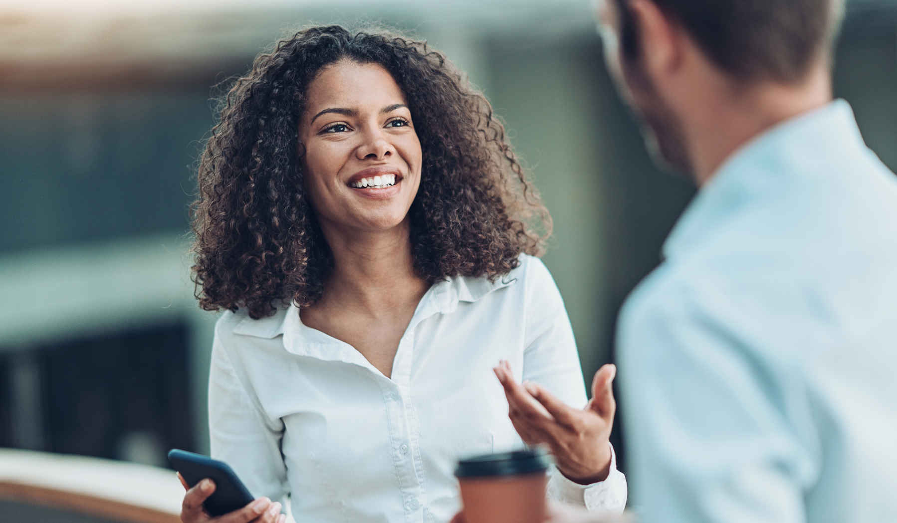 Bild zeigt eine Frau mit Smartphone in weißem Hemd, die in einem Gespräch mit einem Mann in blauem Hemd spricht. Der Mann ist mit Rücken zur Kamera gewandt und hält einen Kaffee-Becher in der Hand. Das Gespräch scheint entspannt zu sein, denn die Frau hat gerade das Liquiditätszertifikat erworben.