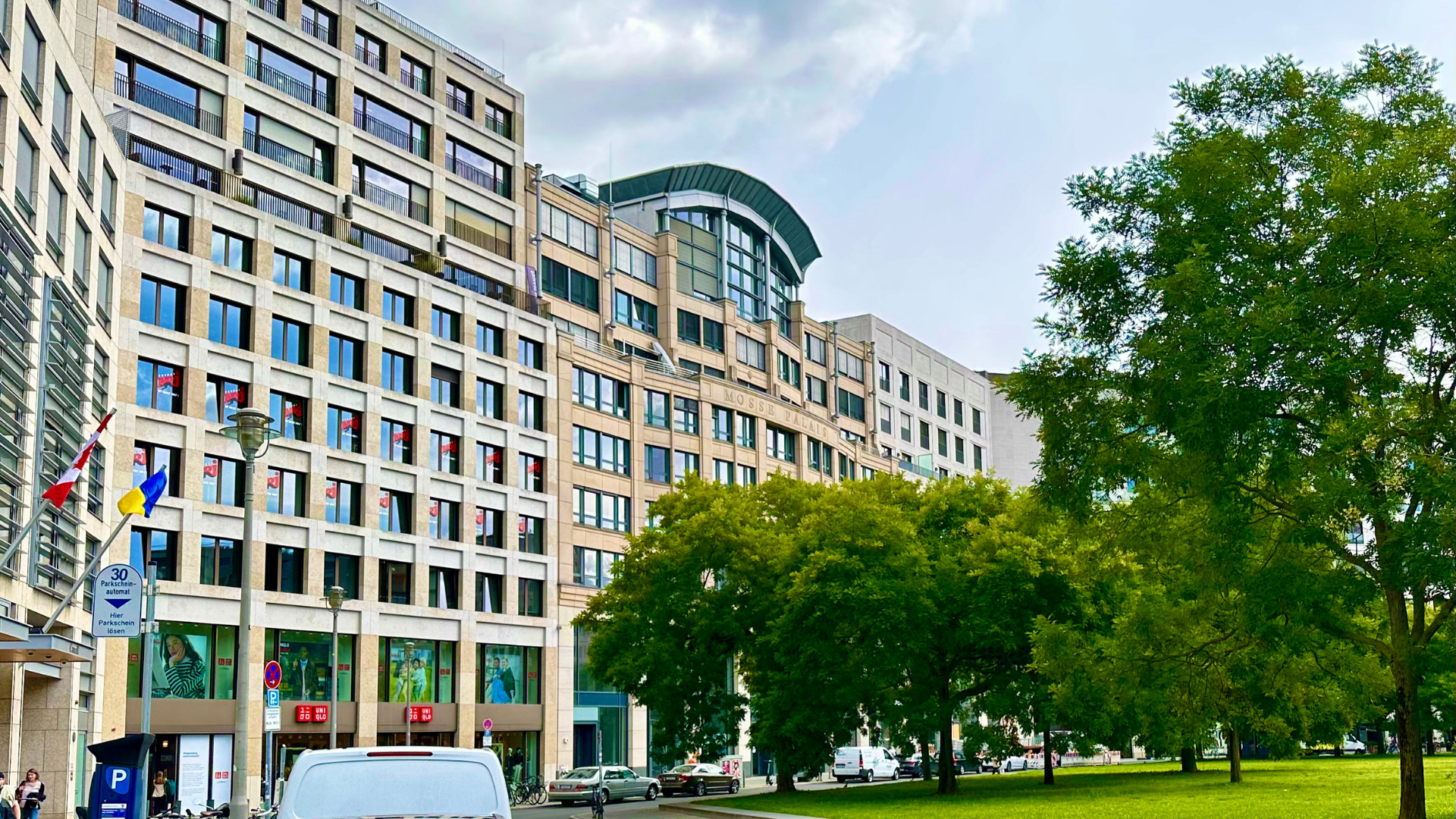 Außenansicht des Mosse Palais am Leipziger Platz in Berlin. Das mehrstöckige Gebäude mit moderner Sandsteinfassade beherbergt mehrere Büros. Im Vordergrund sind Bäume, eine Parkfläche und Fahrzeuge zu sehen, während sich Flaggen und ein Parkautomat am Gehweg befinden.