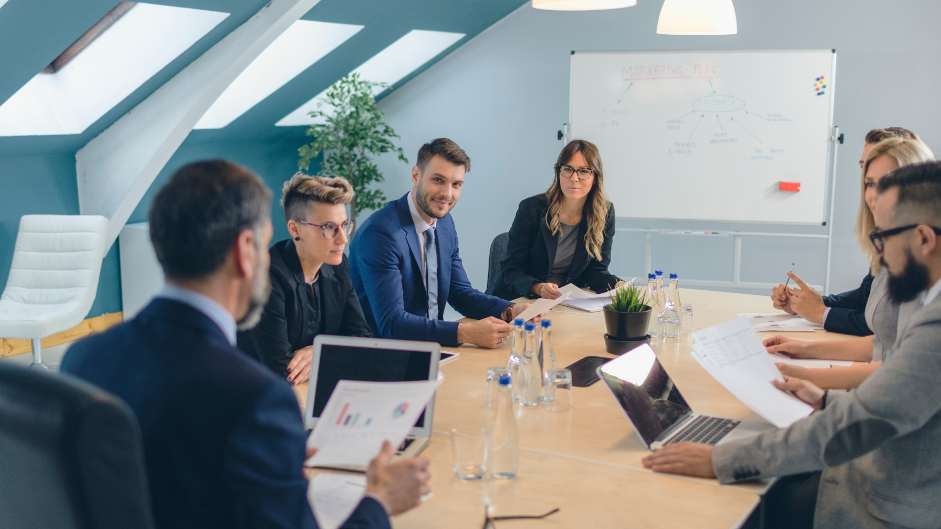 Eine Gruppe von acht Personen in formeller Kleidung sitzt in einem modernen Konferenzraum um einen großen Tisch. Auf dem Whiteboard im Hintergrund sind Begriffe wie „Marketing Plan“ und „Feedback“ zu lesen. Die Teilnehmenden analysieren Dokumente und besprechen Strategiepläne.