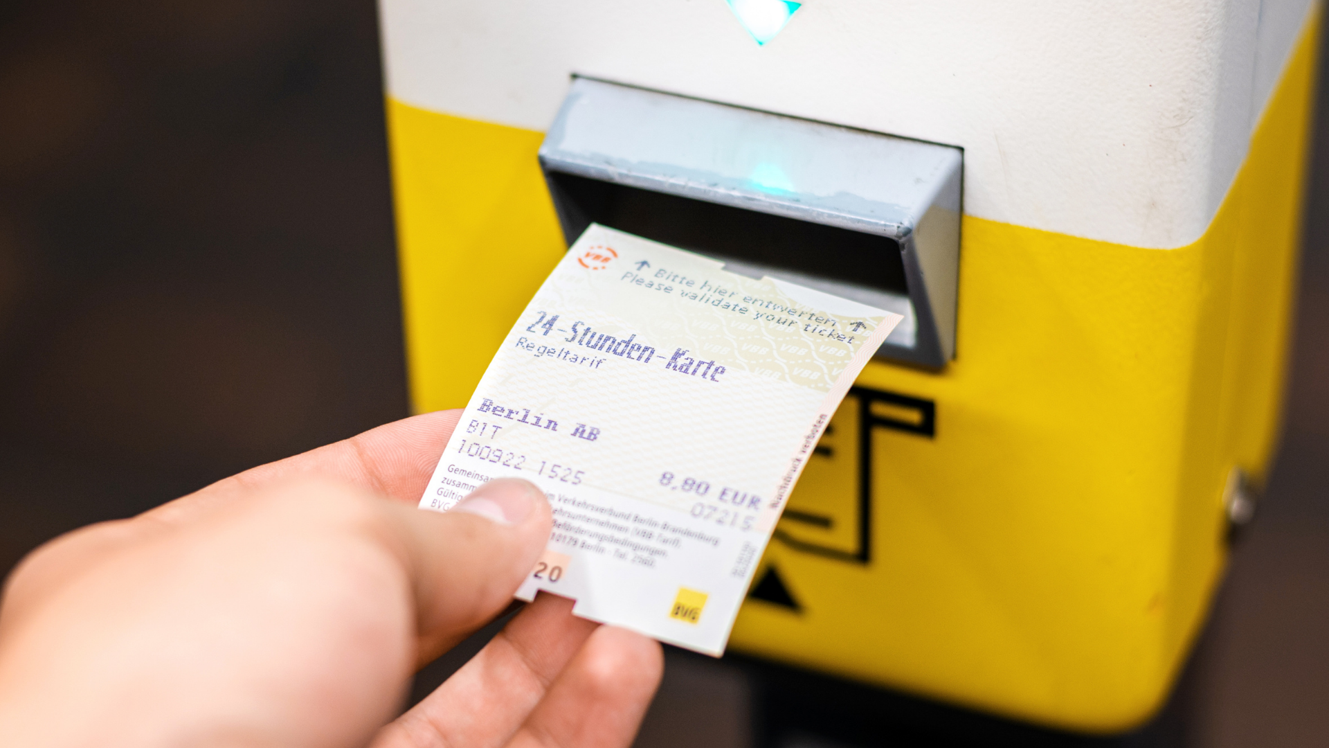 Das Bild zeigt eine Hand, die eine Fahrkarte vor einem gelben Ticket-Entwerter in einem öffentlichen Verkehrsmittel hält. Die Fahrkarte ist für 24 Stunden gültig und spezifisch für die Tarifzone Berlin AB, mit einem Preis von 8,90 Euro. Das Datum auf dem Ticket ist der 10. März 2022, und es ist 15:25 Uhr. Auf dem Ticket steht außerdem der Hinweis, es zu entwerten, was darauf hinweist, dass es vor Beginn der Reise gestempelt werden muss.