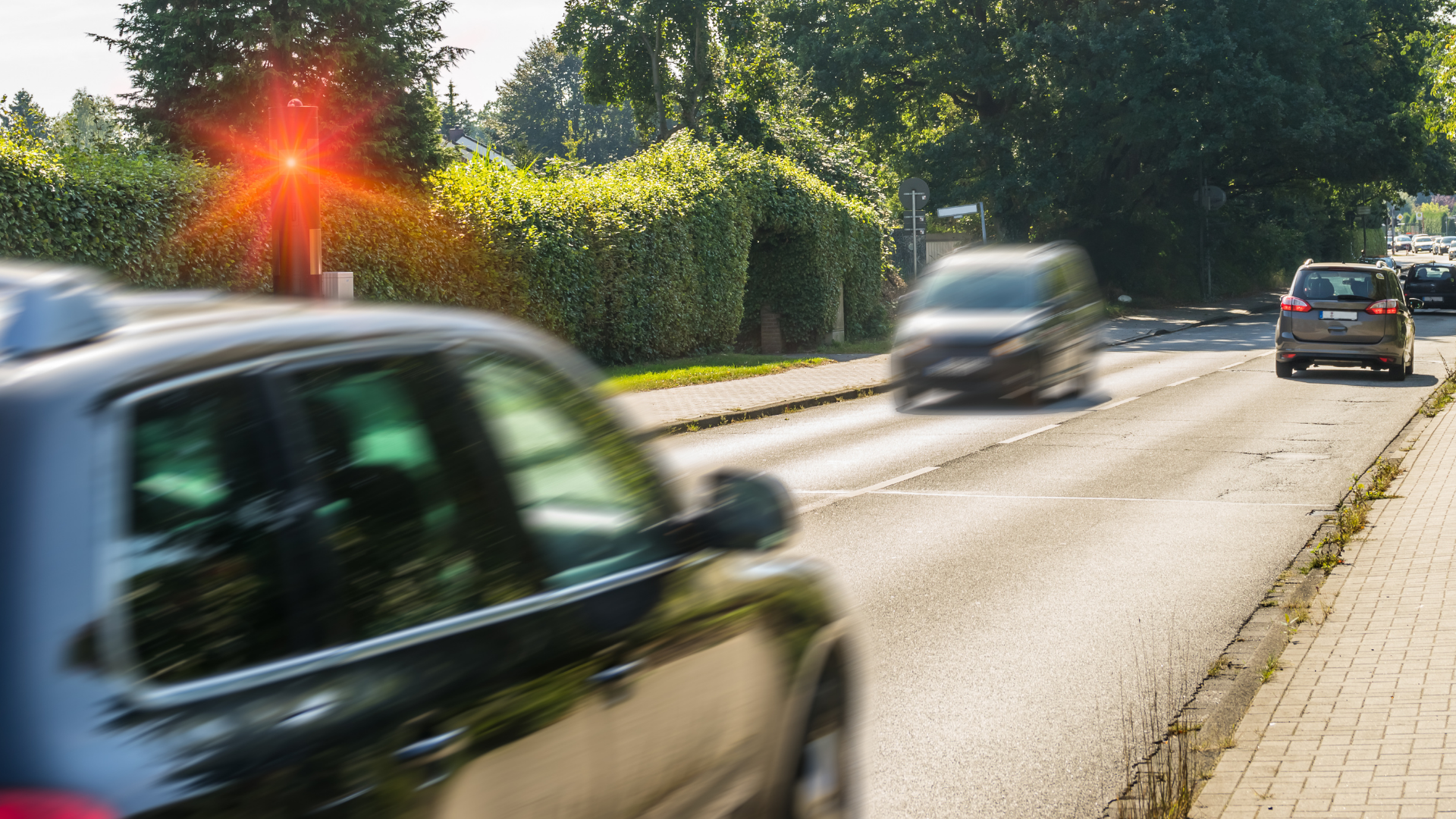 Das Bild zeigt eine städtische Straßenszene, auf der Autos in verschiedenen Geschwindigkeiten fahren. Auffallend ist ein Geschwindigkeitsüberwachungsgerät am Straßenrand, das durch ein rotes Blitzlicht markiert ist, was darauf hinweist, dass es möglicherweise ein Fahrzeug beim Überschreiten der Geschwindigkeitsbegrenzung erfasst hat. Die Szene vermittelt eine alltägliche Verkehrssituation, in der Geschwindigkeitskontrollen dazu beitragen, die Sicherheit auf den Straßen zu erhöhen und Verkehrsteilnehmer zur Einhaltung der Regeln anzuhalten.