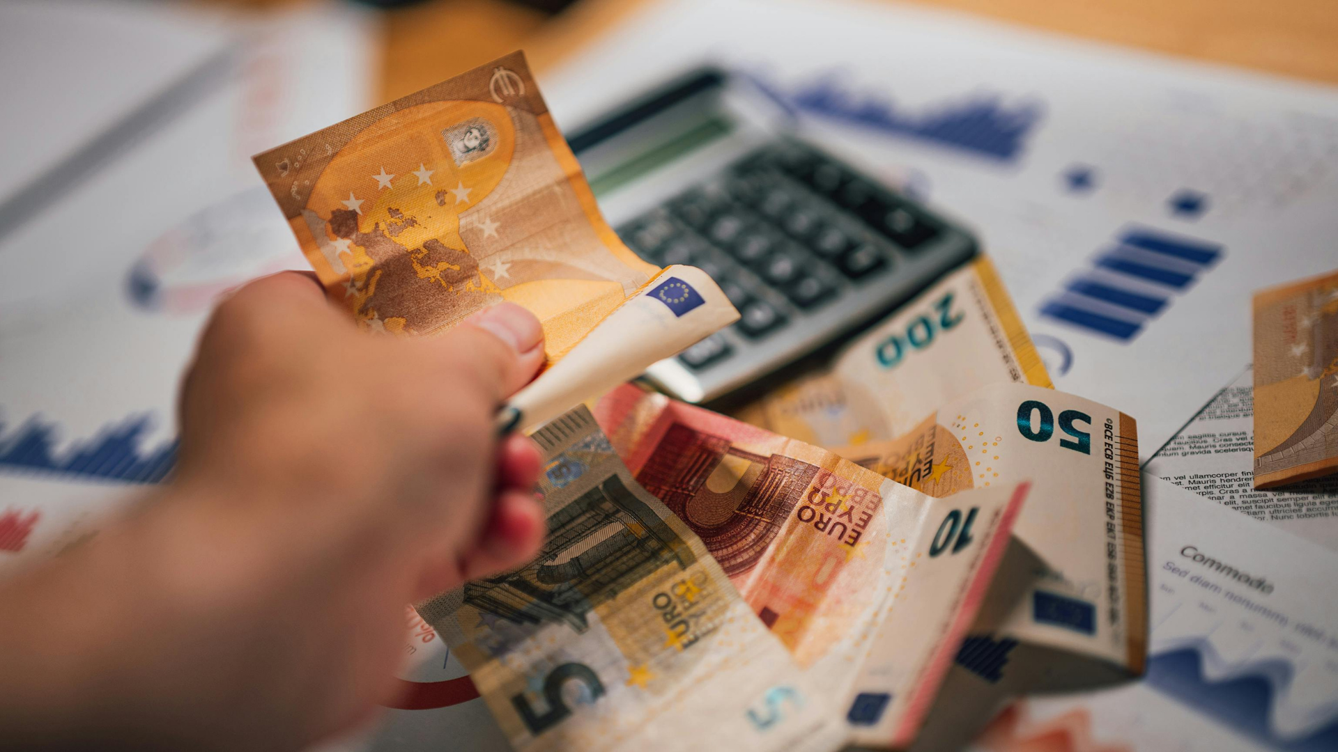 Das Bild zeigt eine Hand, die einen 50-Euro-Schein hält, über einem Tisch, der mit weiteren Euro-Banknoten und einer Taschenrechner bedeckt ist. Im Hintergrund sind Papierdokumente mit Finanzdiagrammen zu sehen. Diese Szene vermittelt das Thema finanzielle Planung oder Buchhaltung, wobei Geldscheine und Finanzdokumente die Analyse und Verwaltung von Finanzen darstellen. Die klare Darstellung von Euro-Banknoten und das Vorhandensein von Finanzmaterialien deuten auf die Durchführung von Budgetberechnungen oder finanziellen Bewertungen hin.